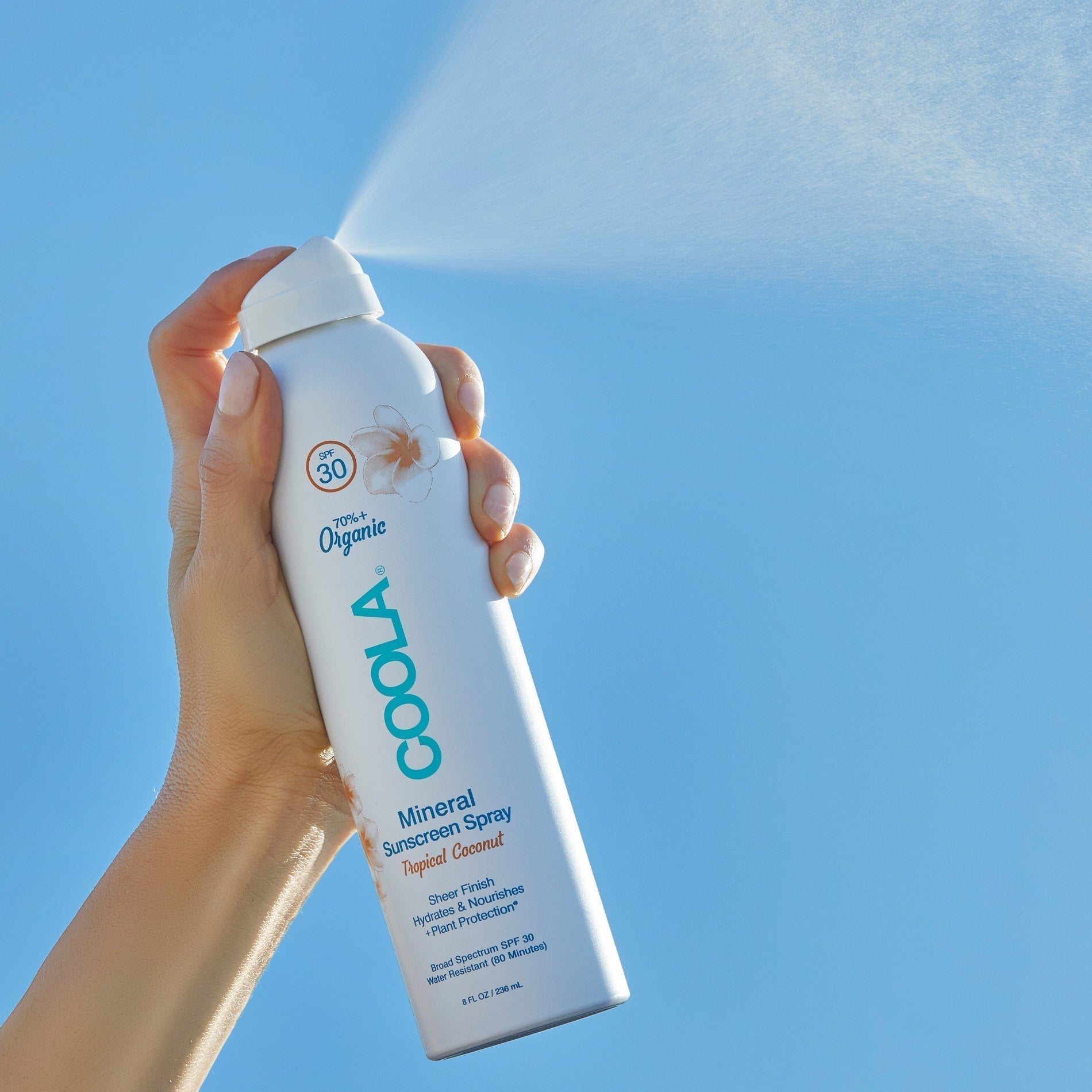 Hand spraying COOLA Mineral Sunscreen Spray SPF 30 Tropical Coconut against a blue sky.
