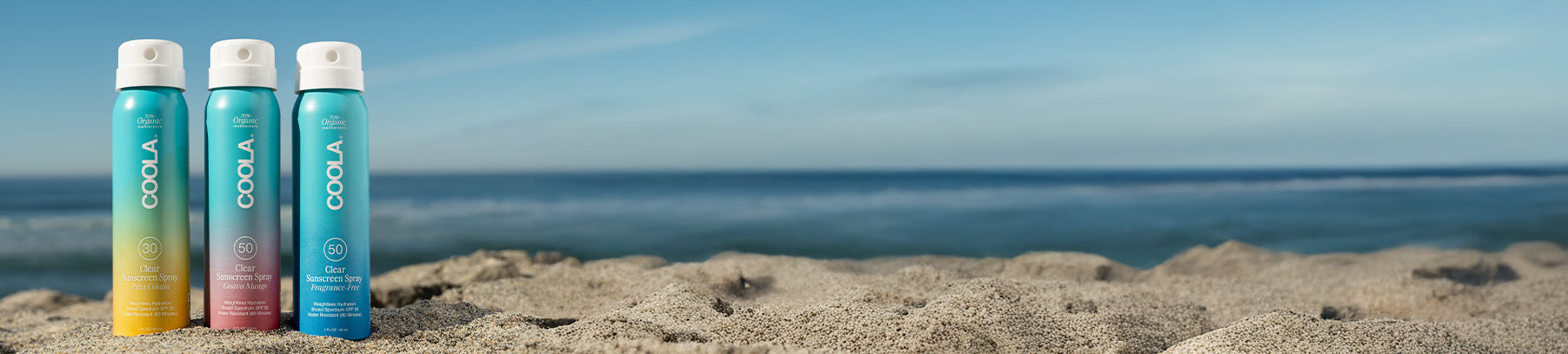 Three COOLA Clear Sunscreen Spray SPF bottles in the sand