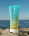 COOLA Hydrating Sunscreen Lotion Piña Colada SPF 30 sitting in sand with scenic ocean background