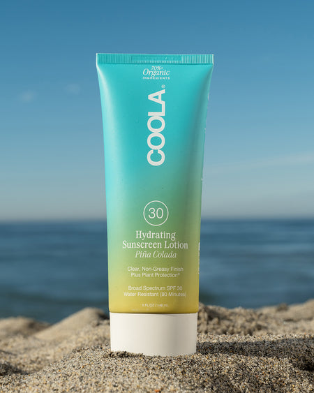 COOLA Hydrating Sunscreen Lotion Piña Colada SPF 30 sitting in sand with scenic ocean background