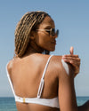 Woman in white bikini applying COOLA body lotion sunscreen to shoulder with scenic ocean background