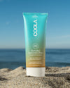 COOLA Hydrating Sunscreen Lotion Tropical Coconut SPF 30 sitting in sand with scenic ocean background