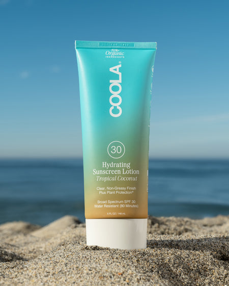 COOLA Hydrating Sunscreen Lotion Tropical Coconut SPF 30 sitting in sand with scenic ocean background