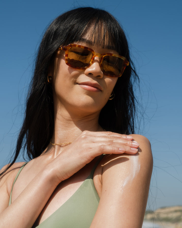 Woman applying COOLA hydrating sunscreen lotion to shoulder with scenic coastal background