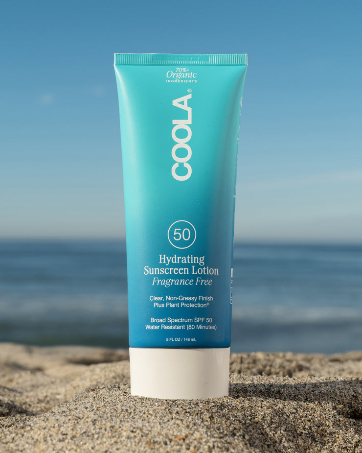 COOLA Hydrating Sunscreen Lotion Fragrance-Free SPF 50 sitting in sand with scenic ocean background