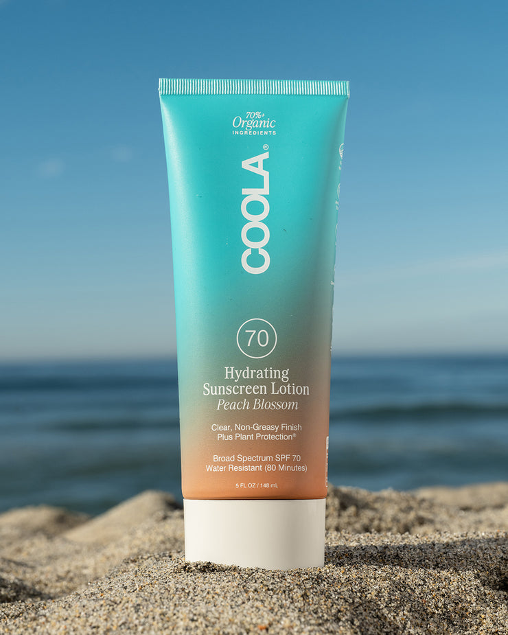 COOLA Hydrating Sunscreen Lotion Peach Blossom SPF 70 sitting in sand with scenic ocean background