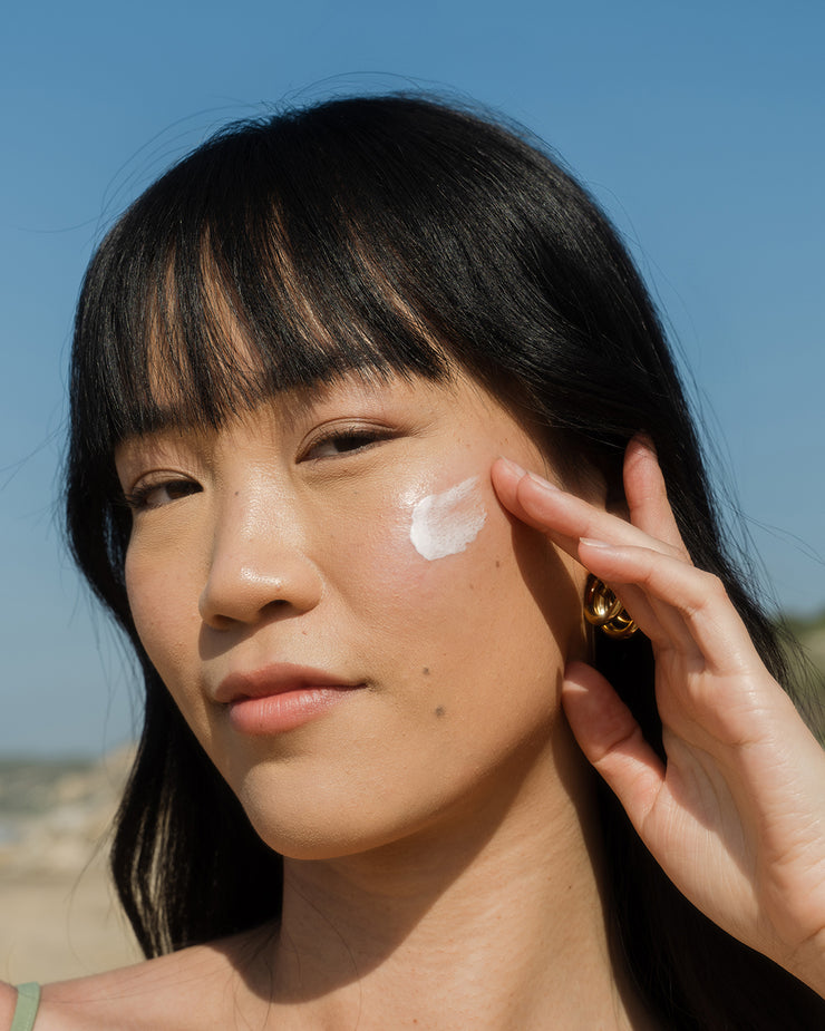 Woman applying COOLA classic face lotion sunscreen to cheek with coastal scenic background