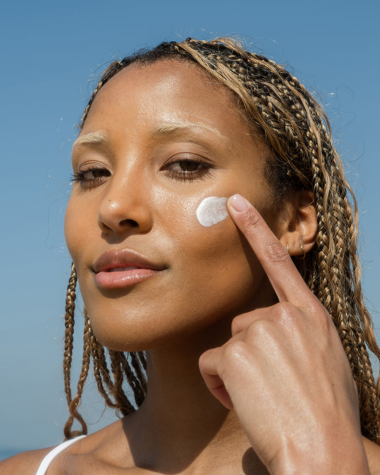 Woman applying COOLA classic face lotion sunscreen to cheek with blue sky background