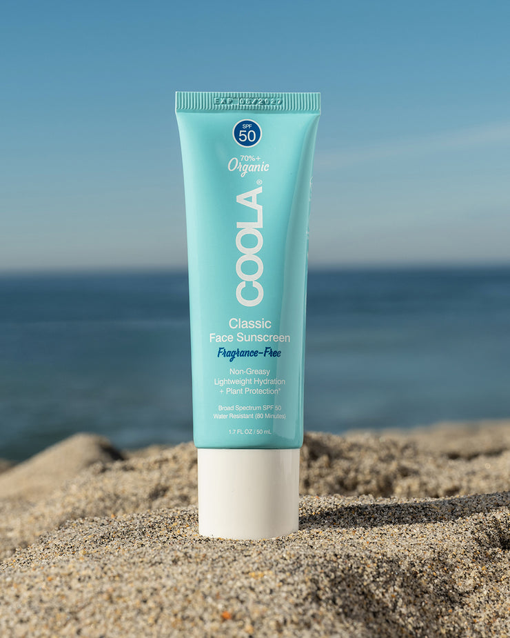COOLA Classic Face Sunscreen Lotion Fragrance-Free SPF 50 sitting in sand with scenic ocean background