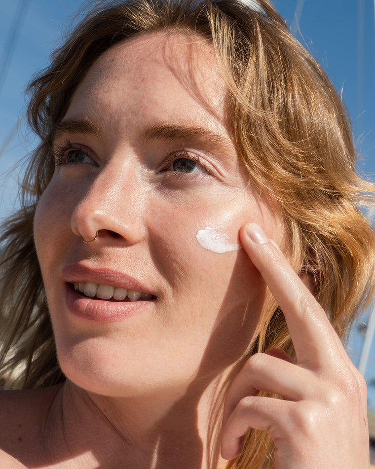 Woman applying COOLA classic face sunscreen lotion to cheek with blue sky background