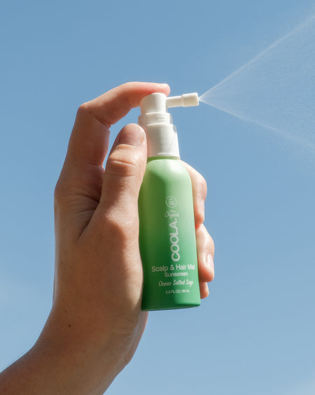 Hand spraying COOLA Scalp & Hair Mist Sunscreen SPF 30 in air against blue sky background