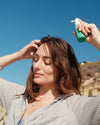 Woman applying COOLA Scalp & Hair Mist Sunscreen SPF 30 to hair against blue sky background