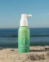 Bottle of COOLA Scalp & Hair Mist Sunscreen SPF 30 sitting in sand with scenic ocean background