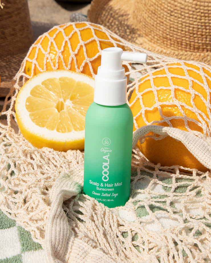 Bottle of COOLA Scalp & Hair Mist Sunscreen SPF 30 sitting on towel with oranges and beach hat in background