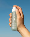 COOLA Makeup Setting Spray being sprayed in air with sky in background