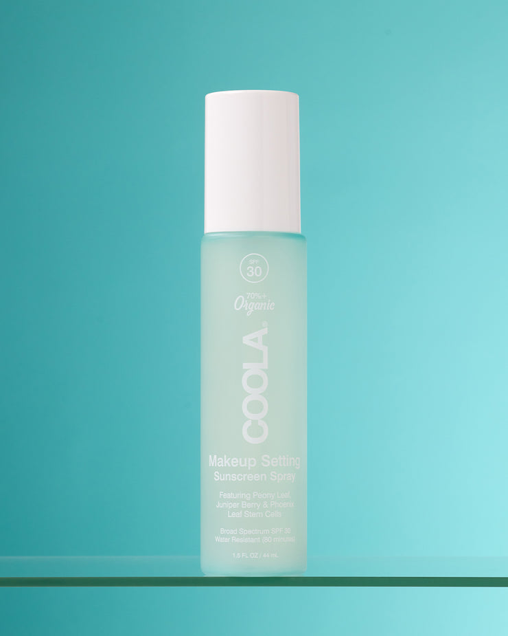 COOLA Makeup Setting Spray sitting on glass shelf against a teal background