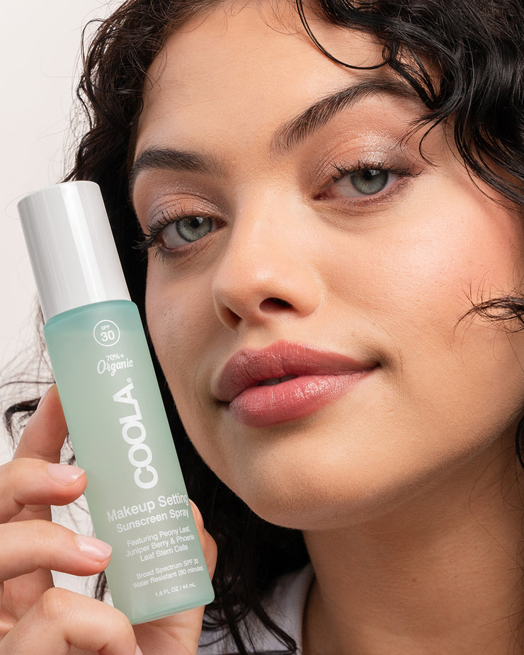 Woman holding COOLA Makeup Setting Spray next to face with white background