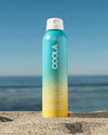 COOLA Clear Sunscreen Spray Piña Colada SPF 30 sitting in sand with scenic ocean background