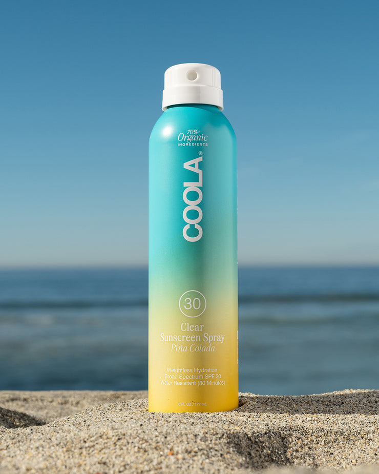 COOLA Clear Sunscreen Spray Piña Colada SPF 30 sitting in sand with scenic ocean background