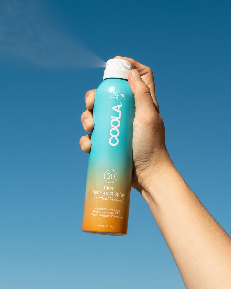 Hand holding COOLA Clear Sunscreen Spray Tropical Coconut SPF 30 spraying in the air with blue sky background