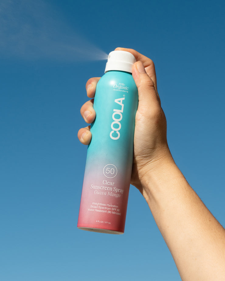 Hand holding COOLA Clear Sunscreen Spray Guava Mango SPF 50 spraying in the air with blue sky background.