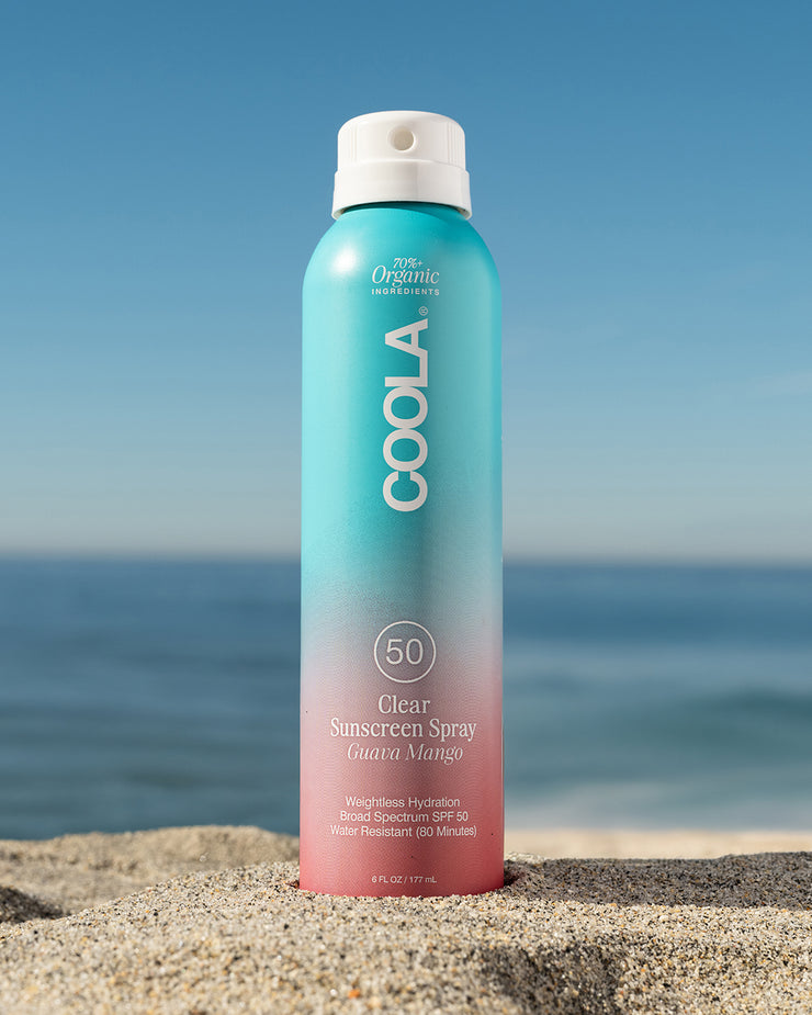 COOLA Clear Sunscreen Spray Guava Mango SPF 50 sitting in sand with scenic ocean background