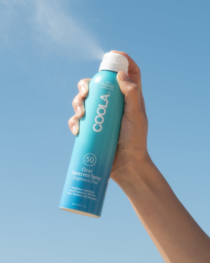 Hand holding COOLA Clear Sunscreen Spray Fragrance-Free SPF 50 spraying in the air with blue sky background