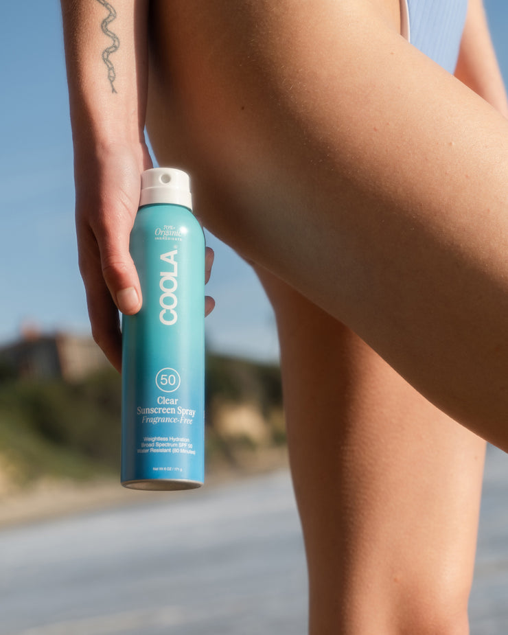 Woman holding COOLA Clear Sunscreen Spray Fragrance-Free SPF 50 next to leg with scenic coastal background