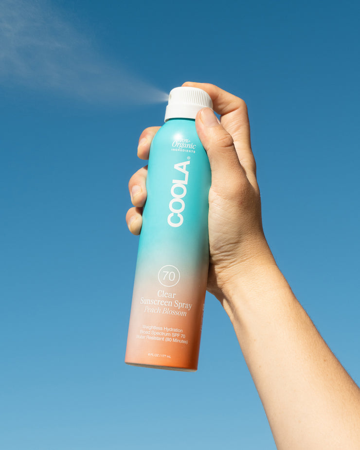 Hand holding COOLA Clear Sunscreen Spray Peach Blossom SPF 70 spraying in air with blue sky background