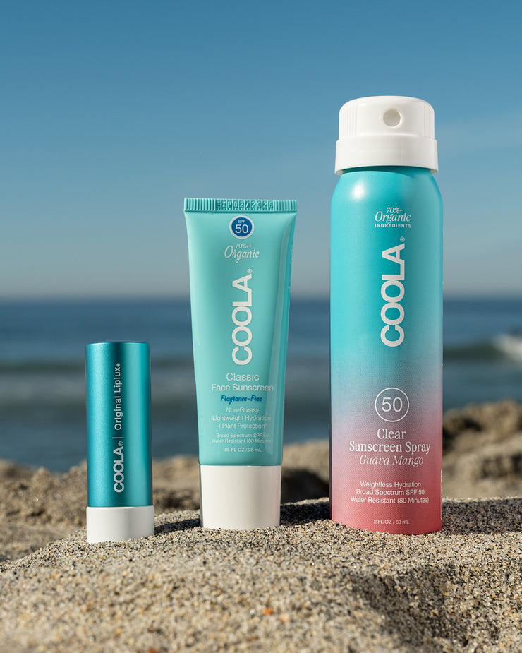 COOLA Classic Liplux, Classic Face Lotion Fragrance-Free, and travel-sized Clear Sunscreen Spray Guava Mango sitting in sand with scenic ocean background.