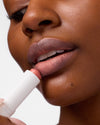 Close-up of woman applying COOLA Nude Beach Mineral Liplux to lips against white background.