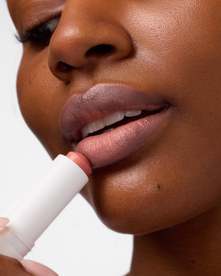 Close-up of woman applying COOLA Nude Beach Mineral Liplux to lips against white background.