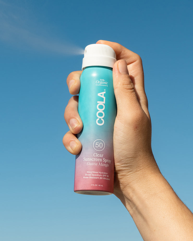Travel-sized COOLA Clear Sunscreen Spray Guava Mango being sprayed with sky blue background.