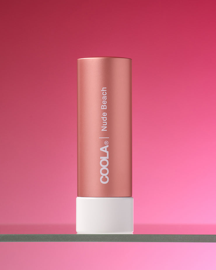 COOLA Nude Beach Mineral Liplux sitting on a glass shelf in peach colored tube against light pink background.