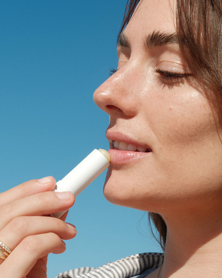 Close-up of woman applying COOLA Classic Liplux against blue sky background.