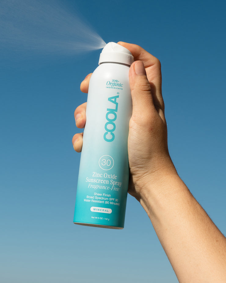 A hand holding a spray bottle of COOLA zinc oxide sunscreen against a blue sky.