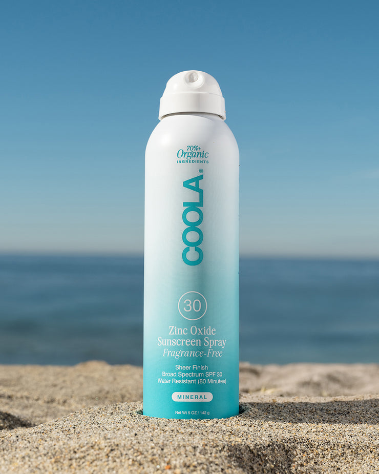 COOLA Zinc Oxide Sunscreen Spray sitting in sand with scenic ocean background