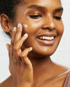 Close-up of woman applying COOLA Vital Rush Skin Renewal Serum with white background.