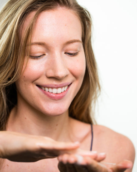 Woman applying COOLA Vital Rush Skin Renewal Serum to hands in bathroom.