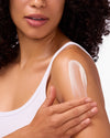 Woman applying COOLA Sunless Tan Firming Lotion to shoulder with white background.