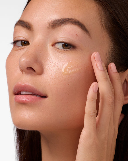 Close-up of woman applying COOLA Sunless Tan Anti-Aging Face Serum with white background.