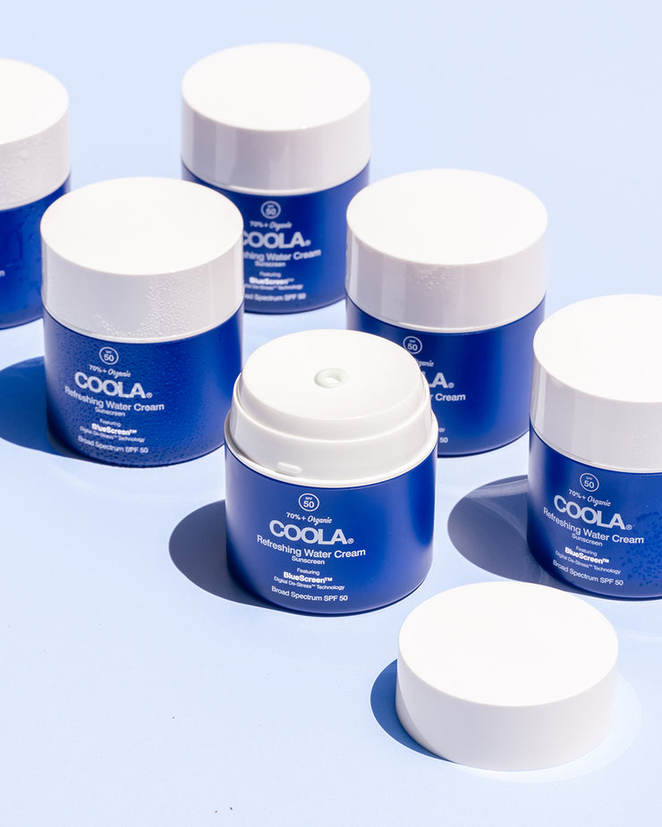 Six COOLA Refreshing Water Cream jars sitting on blue surface