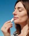 Woman applying COOLA Tinted Liplux to lips with blue sky background