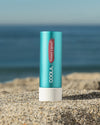 COOLA Tinted Liplux sitting in sand with scenic ocean background