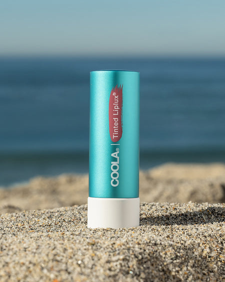 COOLA Tinted Liplux sitting in sand with scenic ocean background