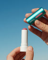 Hands holding COOLA Tinted Liplux being uncapped with blue sky background