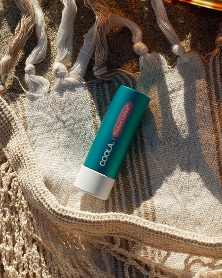 Stick of COOLA Tinted Liplux laying on beach towel