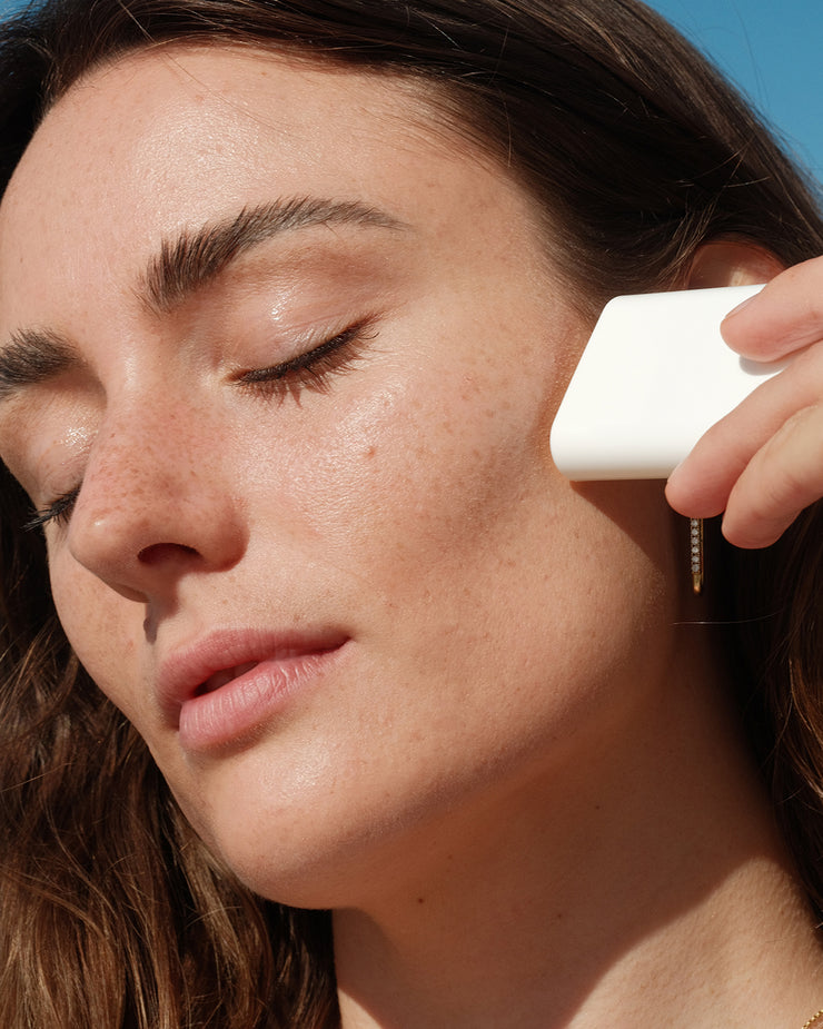 Woman applying COOLA Classic Sunscreen Stick Tropical Coconut to cheek with blue sky background.