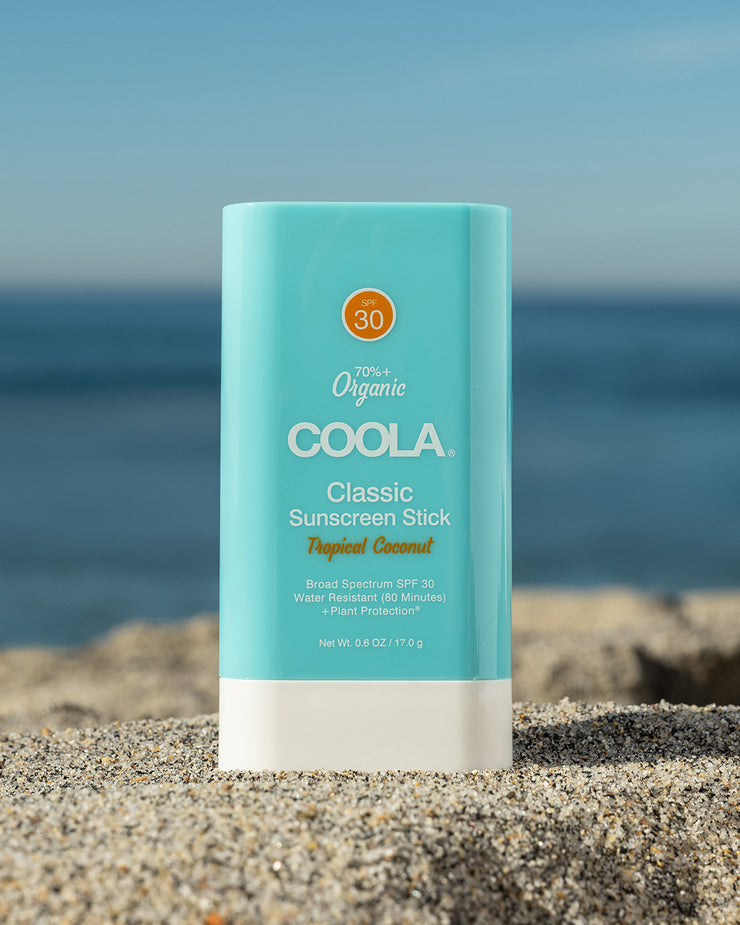 COOLA Classic Sunscreen Stick Tropical Coconut SPF 30 sitting in sand with a scenic ocean background.