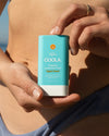 COOLA Classic Sunscreen Stick Tropical Coconut SPF 30 being held in front of woman's body at the beach